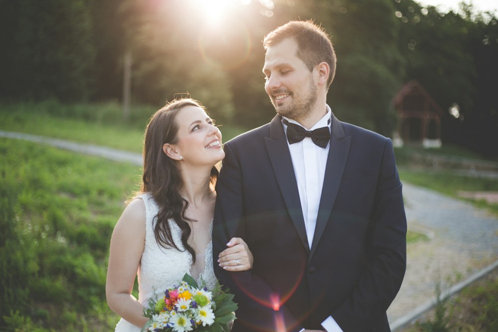 A bride and groom smiling at each other in backlit light beside a forest